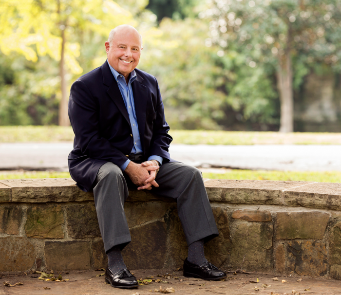A middle-aged man sits casually on a stone bench in a park, wearing a blazer and slacks, with a serene natural background. The setting suggests a relaxed and friendly atmosphere.