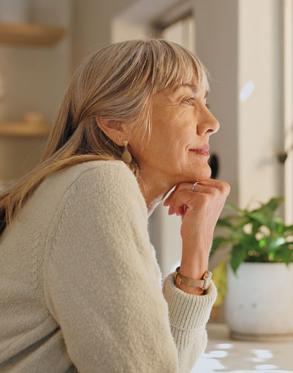 A thoughtful woman with long, gray hair gazes out a window, resting her chin on her hand, while a green plant sits in a pot nearby. Natural light illuminates the scene, creating a calm atmosphere.